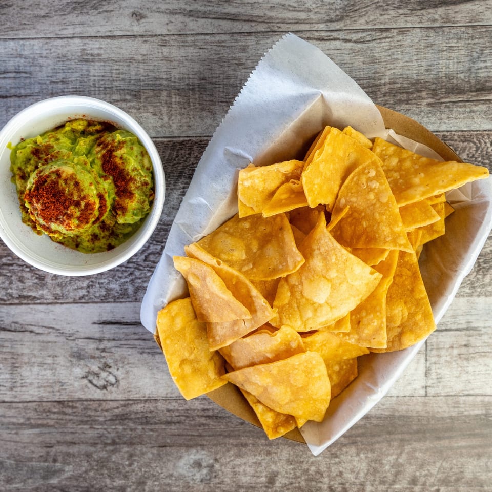 Bowl of seasoned guacamole beside a basket of golden tortilla chips on a rustic wooden table.