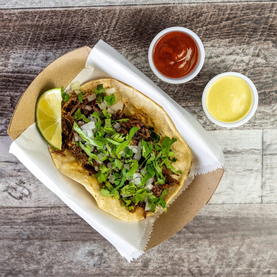 Overhead view of a shredded meat taco topped with onions and cilantro, served with lime and red and yellow sauces.