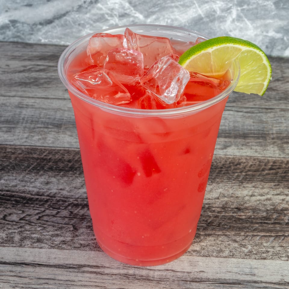 Bright red iced beverage in a clear plastic cup, garnished with a lime slice on a rustic wooden table.