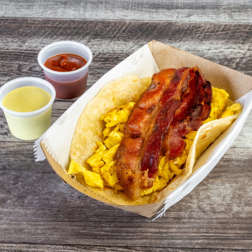 Tortilla wrap filled with scrambled eggs and crispy bacon, served with cups of creamy green and red sauces on a wooden table.