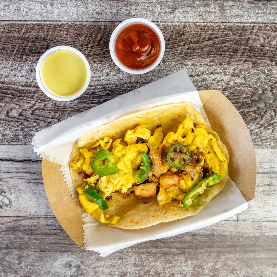 Breakfast burrito filled with scrambled eggs, potatoes, meat, and jalapeños, served with yellow and red sauces on a wooden table.