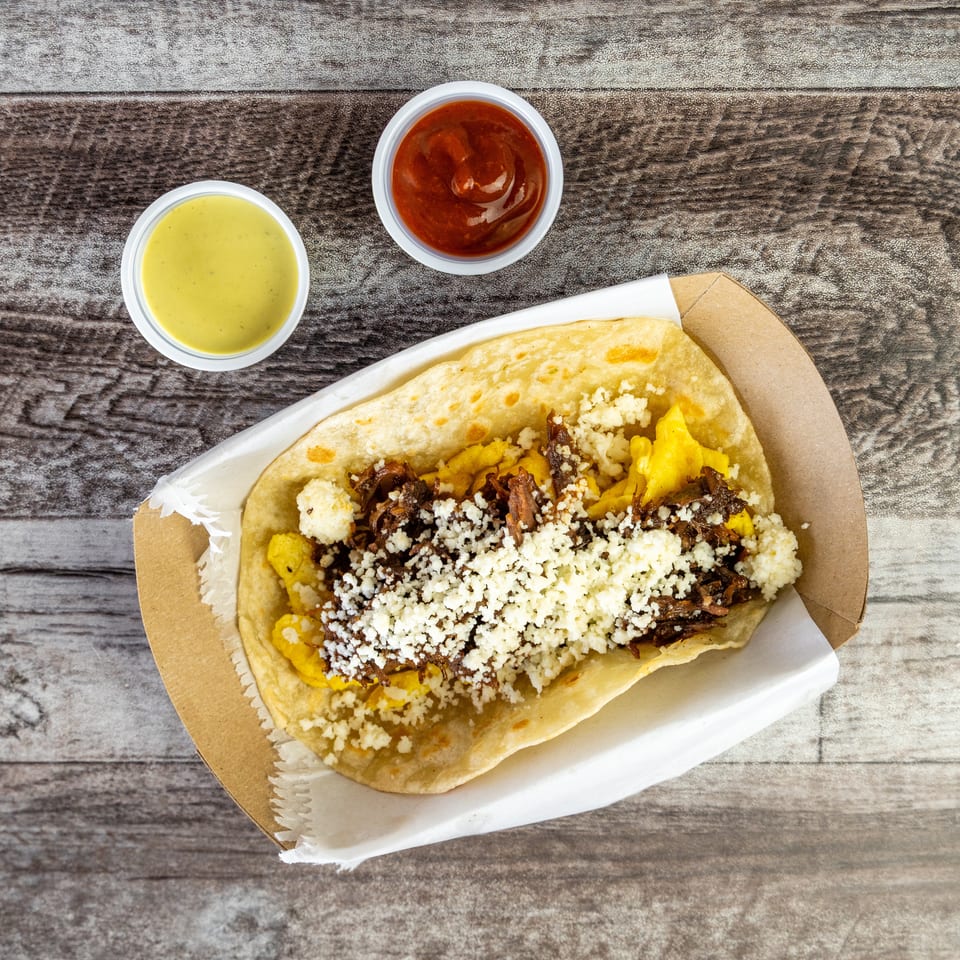 Loaded flatbread topped with shredded meat, scrambled eggs, crumbled cheese, and two dipping sauces on a wooden table.