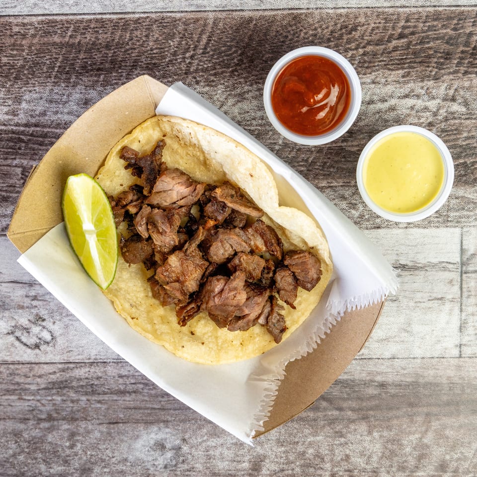 Overhead view of a corn tortilla taco filled with grilled meat, accompanied by a lime wedge and red and yellow sauces.