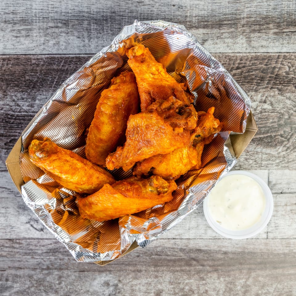 Golden fried chicken wings in a foil-lined basket with a cup of creamy dipping sauce on a wooden table.