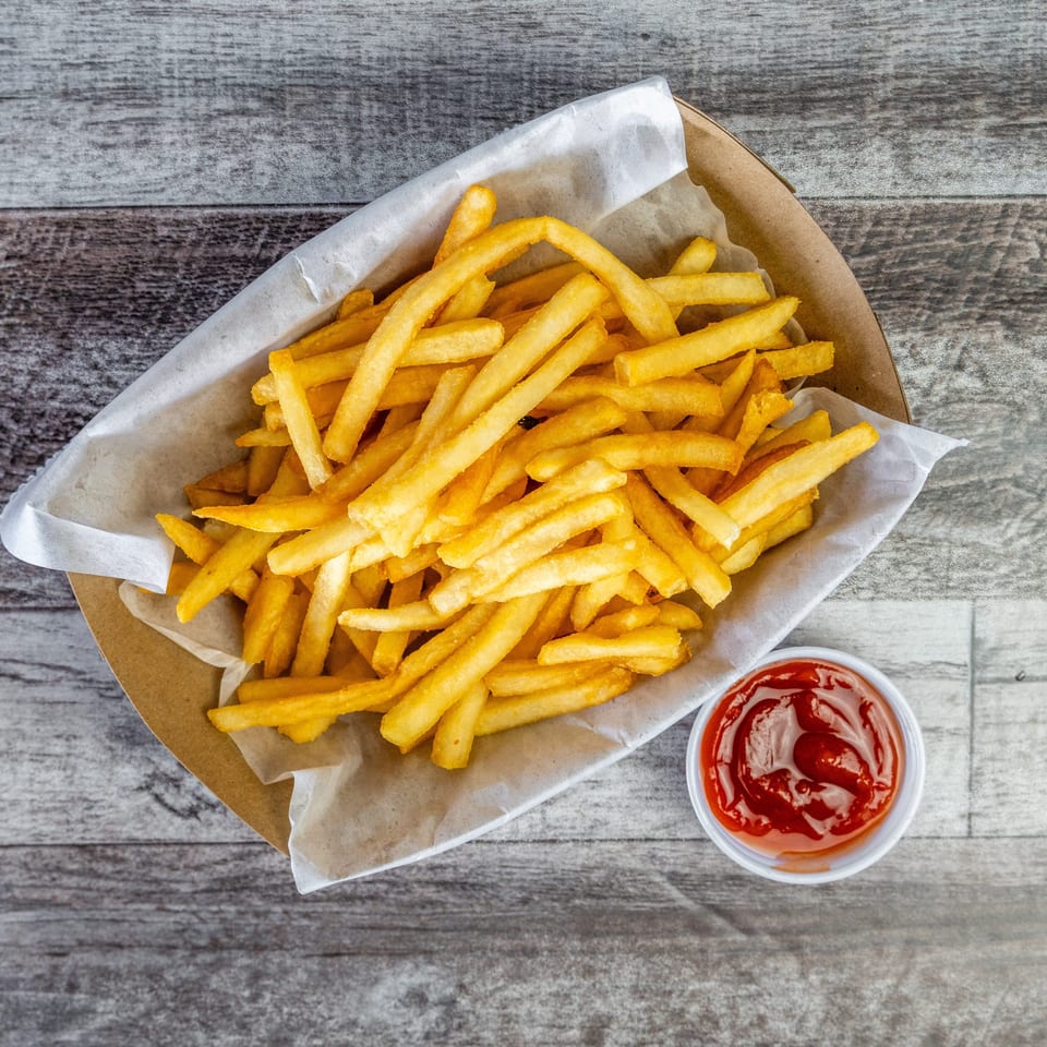 Golden French fries in a paper-lined basket with a cup of ketchup on a rustic wooden table.