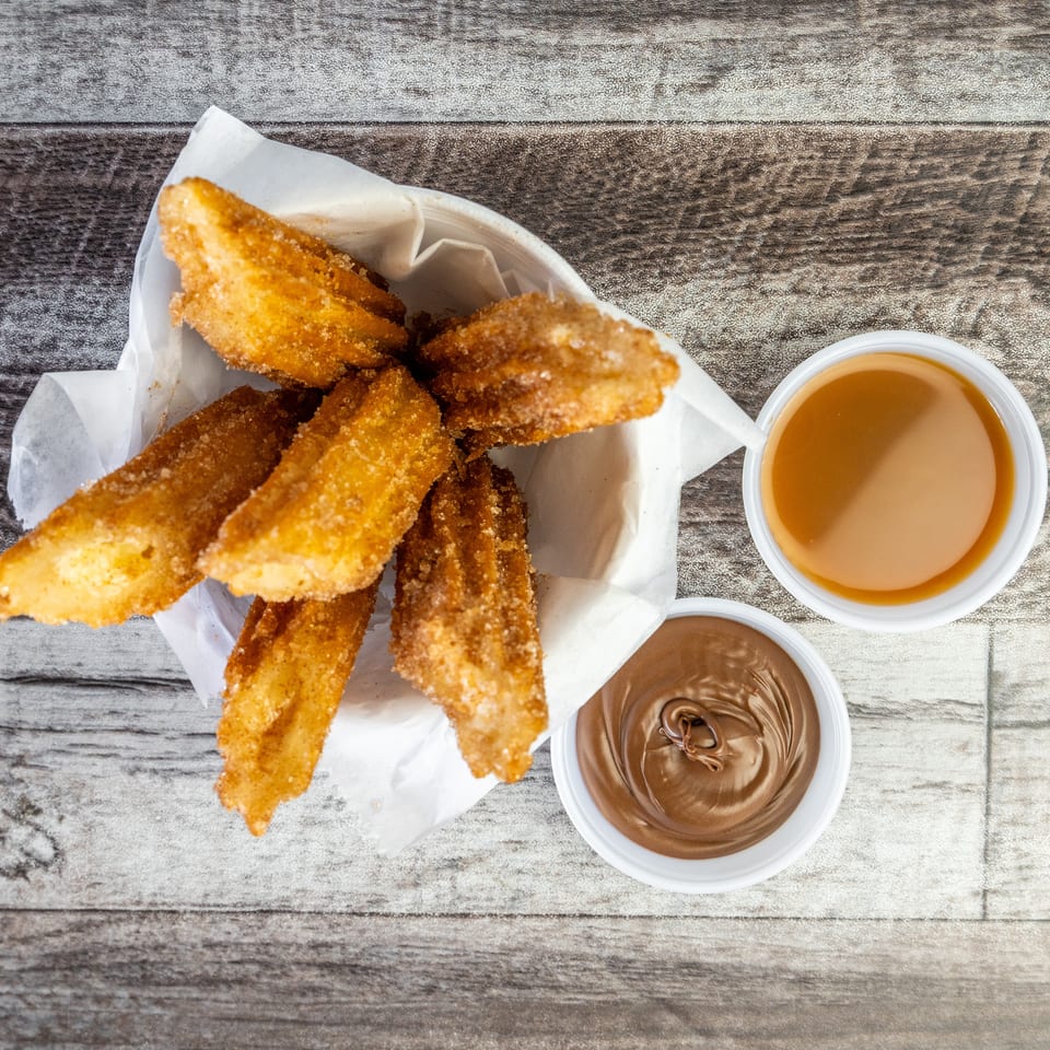 Crispy fried dessert sticks served in a paper-lined basket with caramel and chocolate dipping sauces on a wooden table.