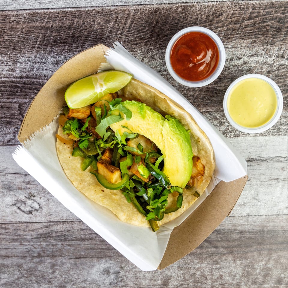 Overhead view of a loaded taco with avocado, roasted vegetables, cilantro, lime, and two dipping sauces on a wooden table.