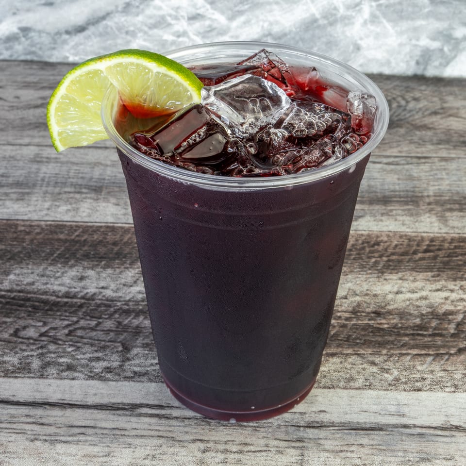 Dark red iced beverage in a clear plastic cup, garnished with a lime wedge on a rustic wooden table.