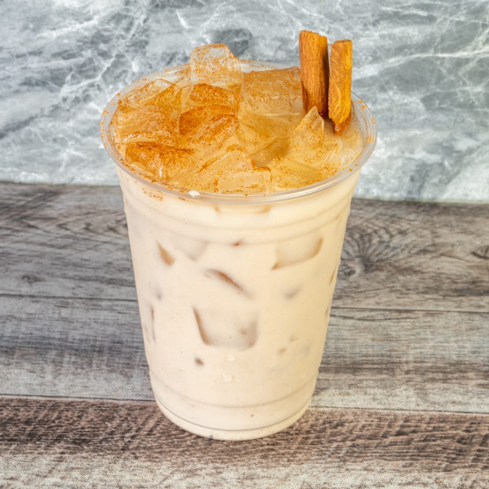 Creamy iced cinnamon drink in a clear cup, topped with ground cinnamon and two cinnamon sticks on a wooden table.