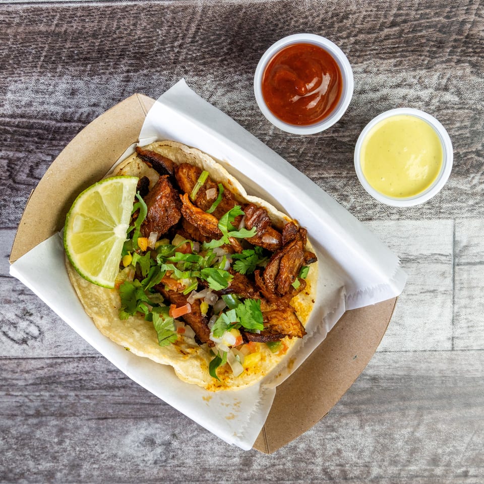 Overhead view of grilled meat tacos topped with cilantro, onions, vegetables, and lime, served with red and yellow sauces.