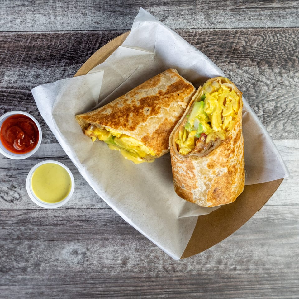 Grilled breakfast burrito cut open to show scrambled eggs, avocado, cheese, and bacon, served with red and green sauces.
