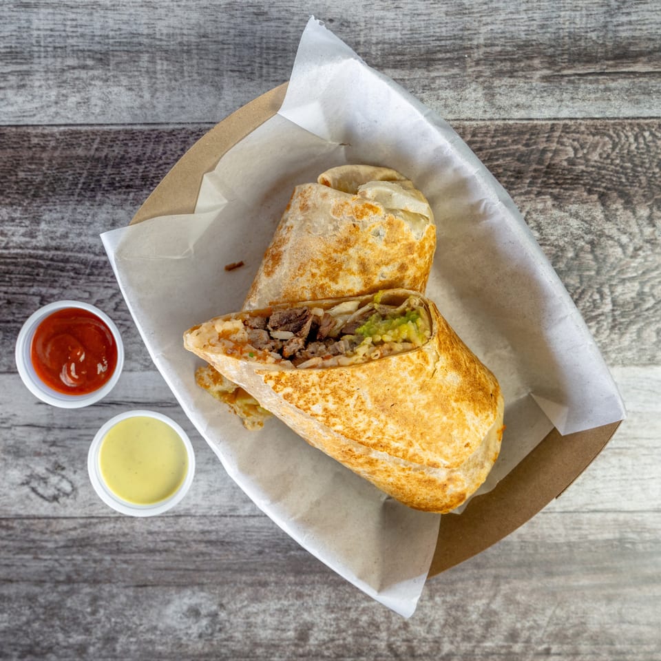 Grilled burrito cut open to reveal steak, rice, and guacamole, served with red and creamy dipping sauces in a paper-lined tray.
