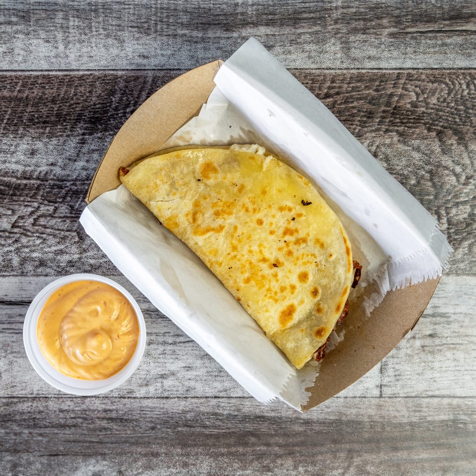 Golden-brown folded quesadilla served in a paper tray with creamy dipping sauce on a rustic wooden table.