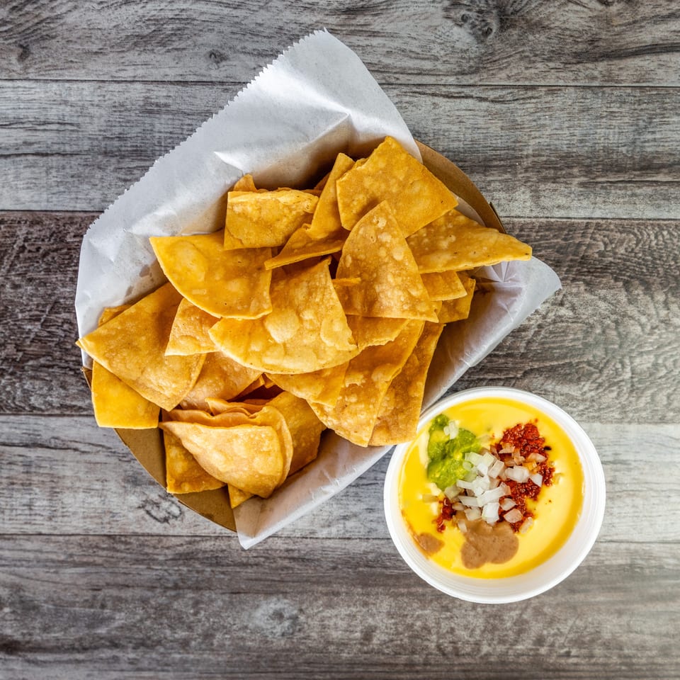 Basket of golden tortilla chips with creamy cheese dip, guacamole, onions, and red salsa on a rustic wooden table.