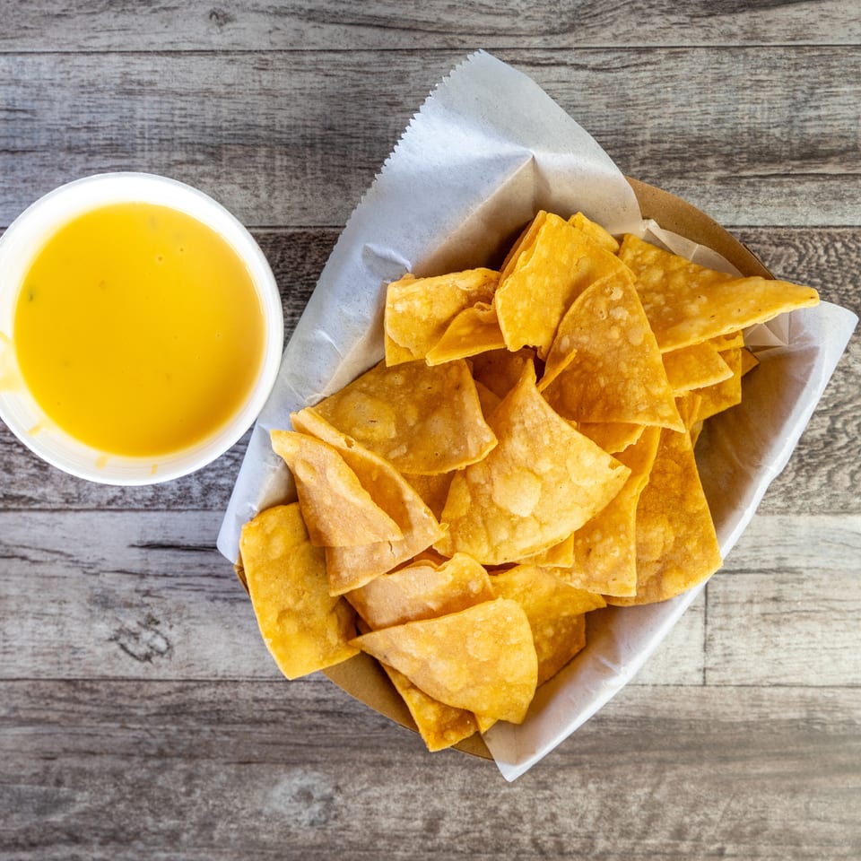 Crispy golden tortilla chips in a paper-lined basket beside a cup of yellow dipping sauce on a rustic wooden table.