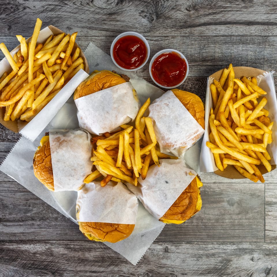 Overhead view of wrapped cheeseburgers, baskets of golden fries, and ketchup cups arranged on a rustic wooden table.