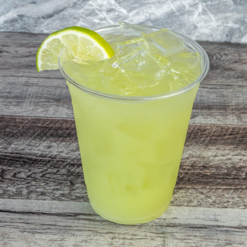 Lime-colored iced drink in a clear plastic cup, garnished with a lime wedge, sits on a rustic wooden table.