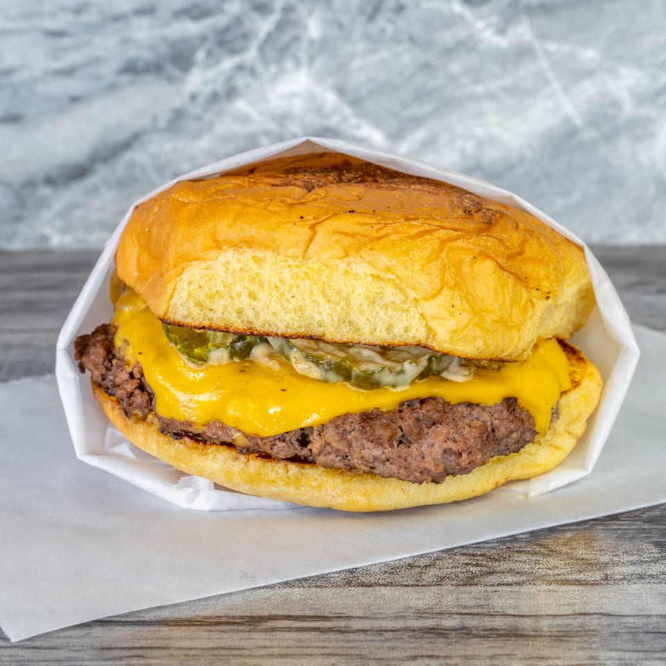 Double cheeseburger with grilled beef patties, melted cheddar, pickles, and sauce on a toasted bun, served in paper wrapping.
